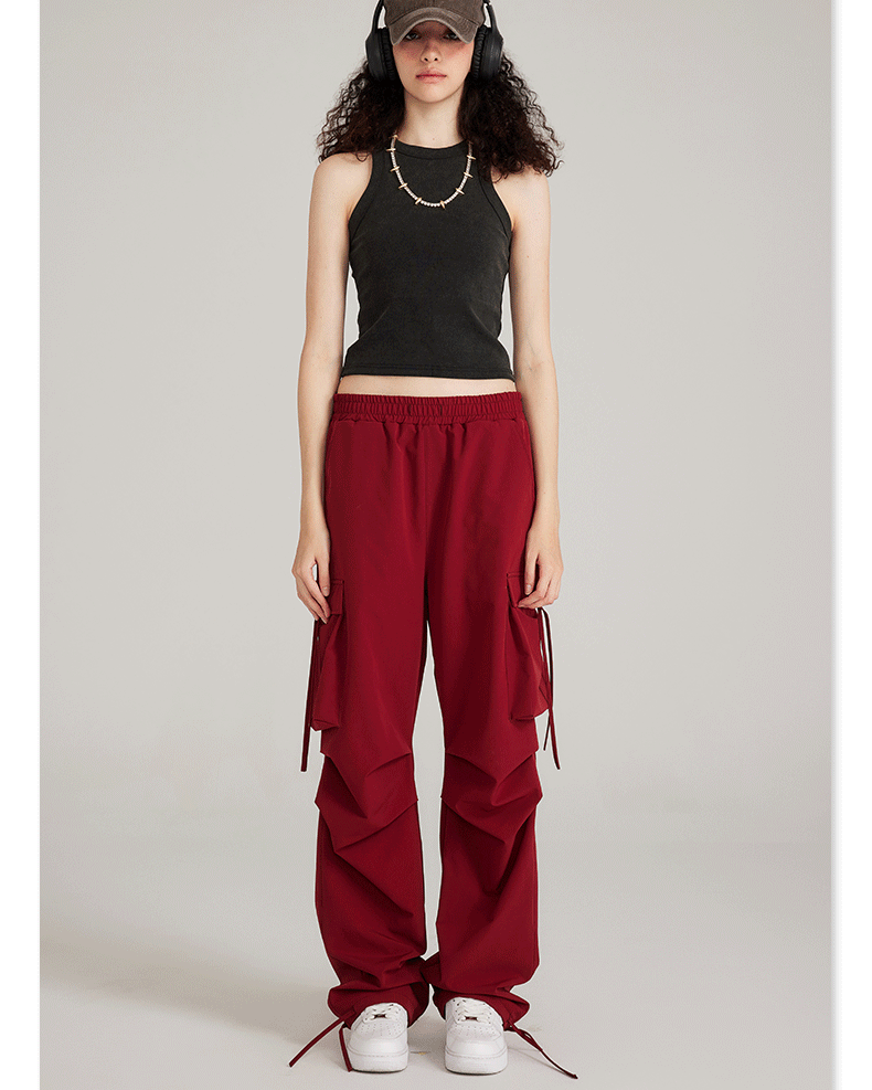 Model wearing black tank top and burgundy cargo pants with side pockets and drawstring details. Styled with cap, headphones and white sneakers