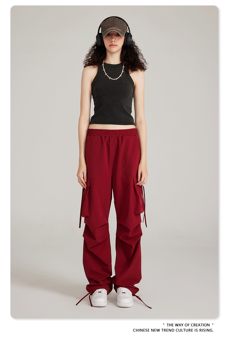 Model wearing black tank top and burgundy cargo pants with side pockets and drawstring details. Styled with cap, headphones and white sneakers
