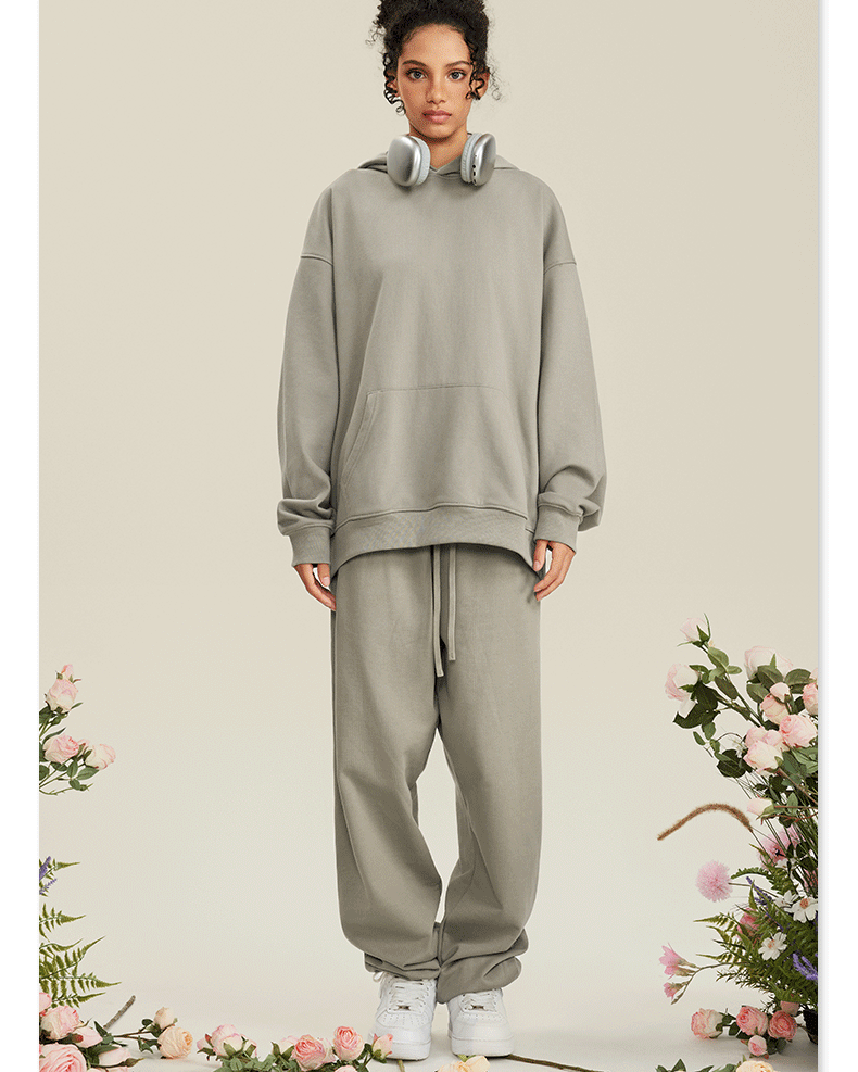 Woman in light gray hoodie and matching joggers with white headphones around neck against floral backdrop