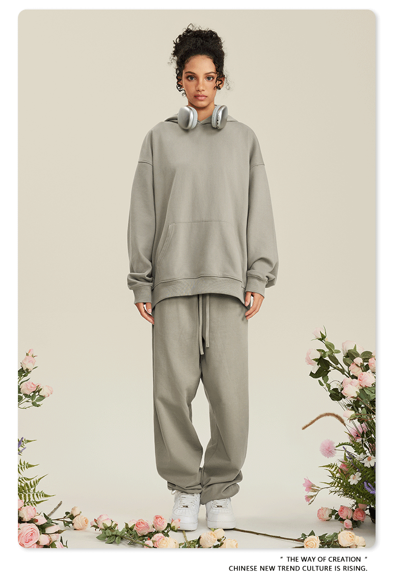 Woman in light gray hoodie and matching joggers with white headphones around neck against floral backdrop