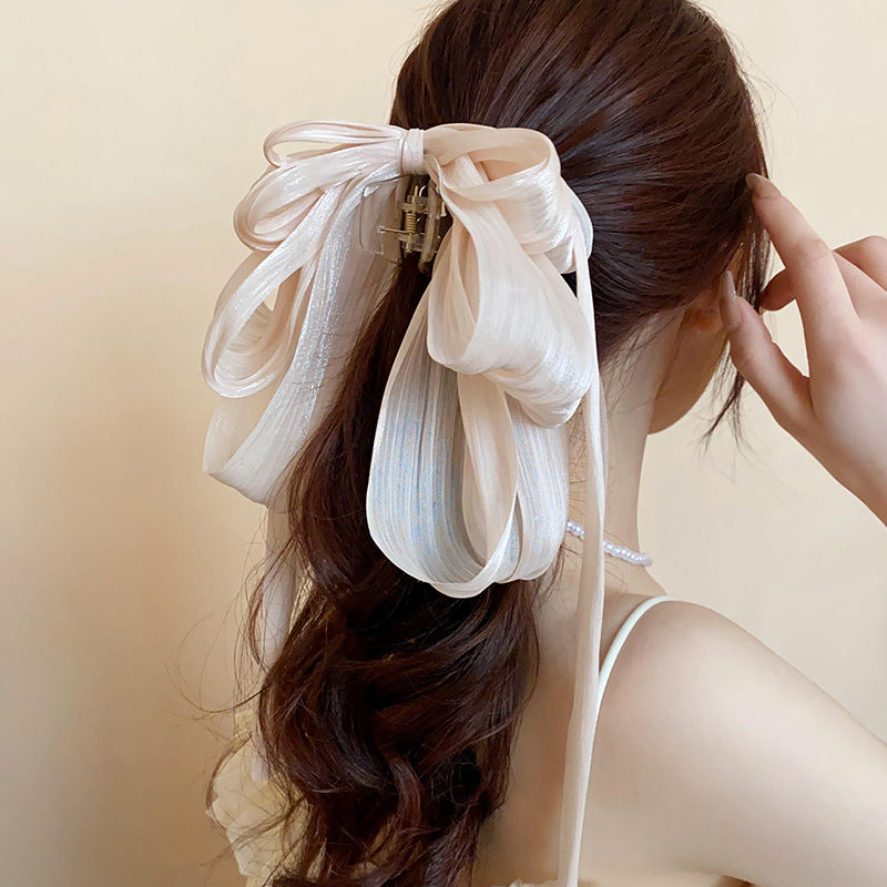 A woman with long brown hair wears a large champagne-colored sheer ribbon bow hair clip, styled in a half-up hairstyle.