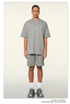 A man with braided hair wearing a light gray oversized t-shirt and shorts set, styled with silver pendant necklace, white socks, and fashion sneakers