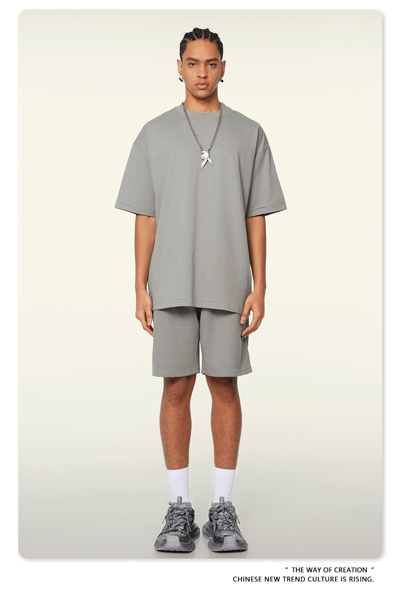 A man with braided hair wearing a light gray oversized t-shirt and shorts set, styled with silver pendant necklace, white socks, and fashion sneakers