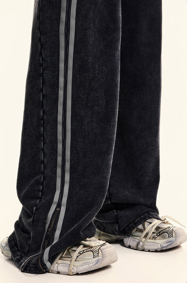 Bottom hem detail of black denim wide-leg pants showing ankle zipper closure with gray stripe trim and athletic footwearClose-up of elastic waistband with drawstring on black washed denim track pants featuring double gray side stripe detail