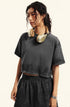 A woman with curly hair wearing gold headphones poses in a dark gray crop top and matching shorts against white backdrop