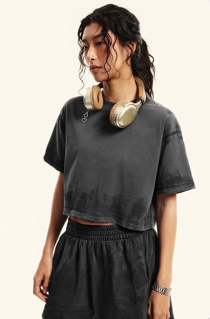A woman with curly hair wearing gold headphones poses in a dark gray crop top and matching shorts against white backdrop