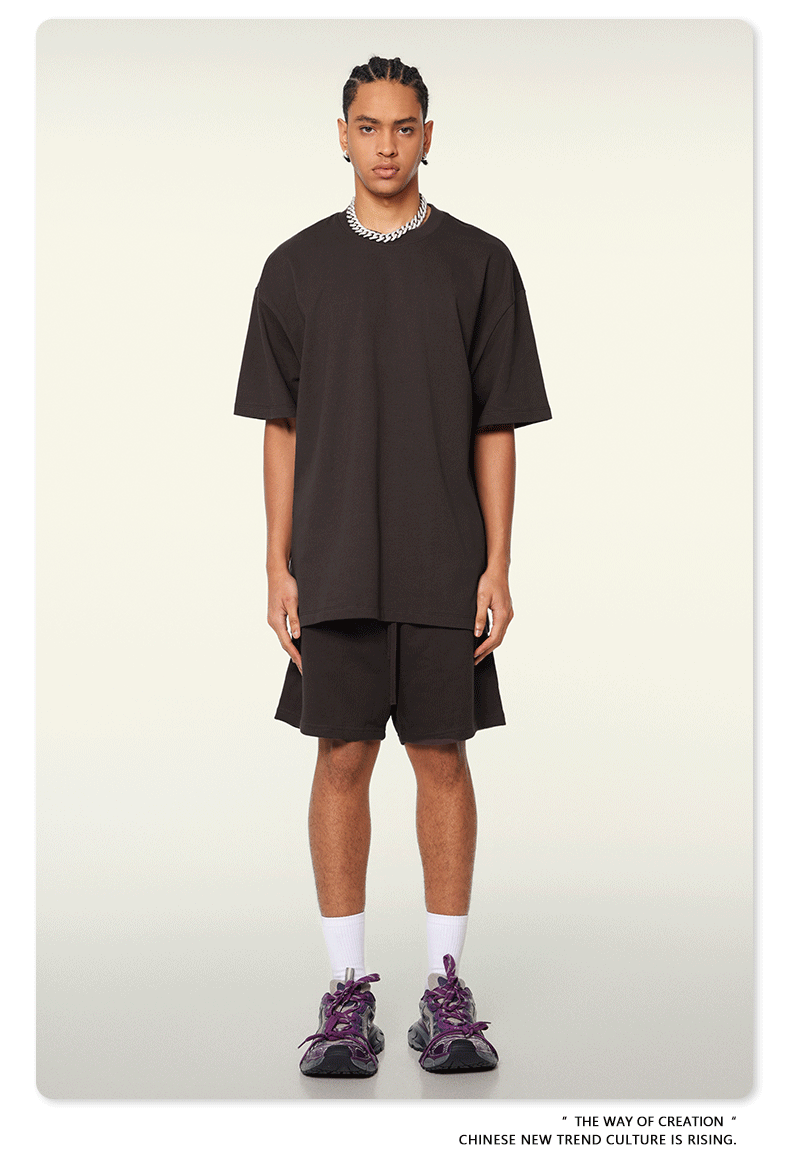 Model with braided hair wearing oversized black t-shirt and matching shorts with white socks and athletic shoes against cream background