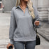 Woman with blonde hair wearing gray textured half-zip pullover with geometric pattern and gold pendant necklace against concrete wall