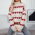 A woman wearing cream ribbed turtleneck sweater with rows of red heart patterns sitting against white wall background holding cream handbag