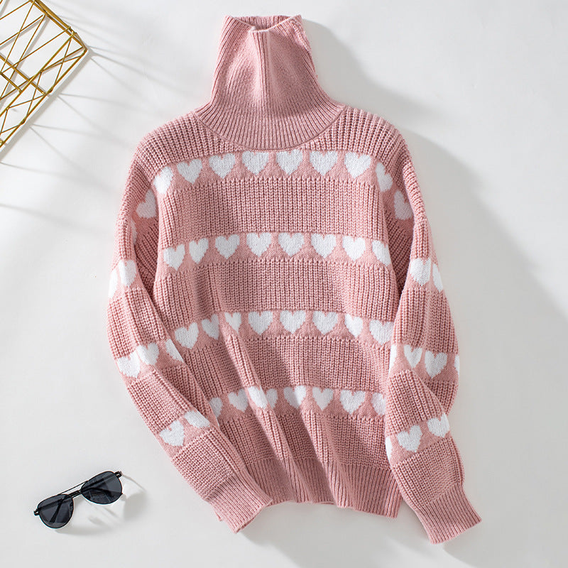 Pink heart pattern turtleneck sweater displayed flat against white background with gold geometric decor and sunglasses visible in corner