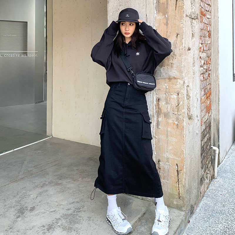 Black cargo maxi skirt with side pockets, styled with Adidas sweatshirt and crossbody bag against industrial backdrop