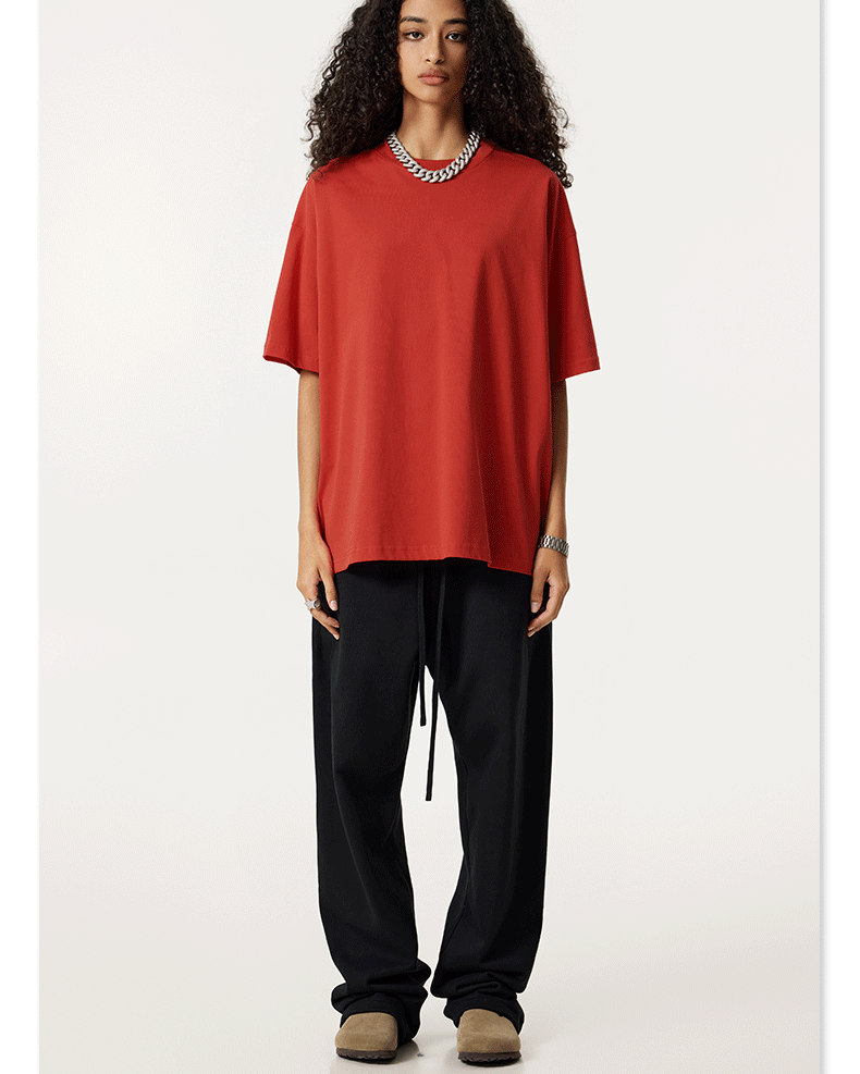 A model with long curly dark hair wearing a coral red oversized t-shirt and black joggers, standing against a white background