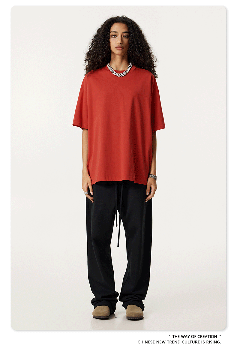 A model with long curly dark hair wearing a coral red oversized t-shirt and black joggers, standing against a white background