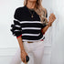 Woman wearing black knit sweater with white stripes and red trim, styled with white jeans and gold chain necklaces in casual winter outfit