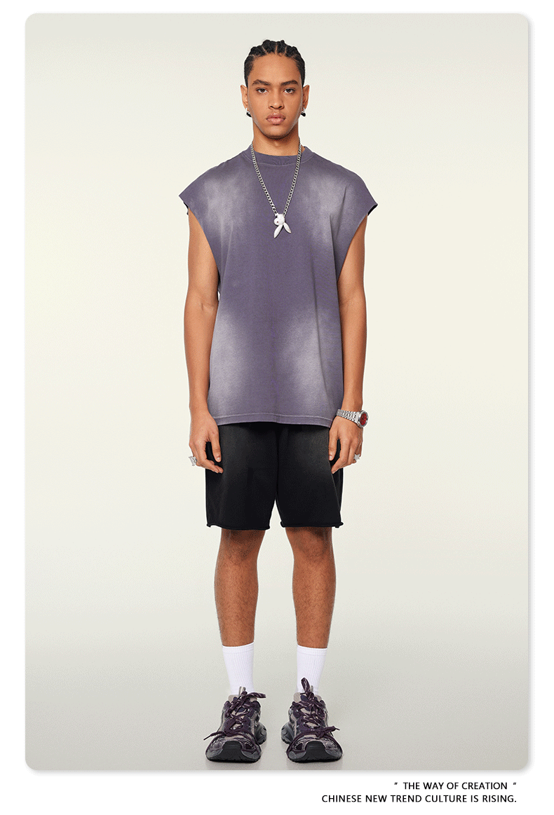 A man wearing a slate blue sleeveless muscle tee and black shorts stands against a white background with silver pendant necklace