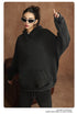 Woman in black oversized hoodie wearing white-framed sunglasses with one hand raised against brown backdrop