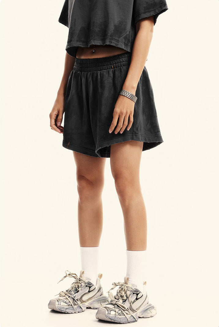 A woman with hand at side wearing dark gray cropped t-shirt, matching shorts, white socks and sneakers against white background