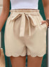 A model wearing beige paperbag shorts with scalloped edges and tie belt, showing front view against green background