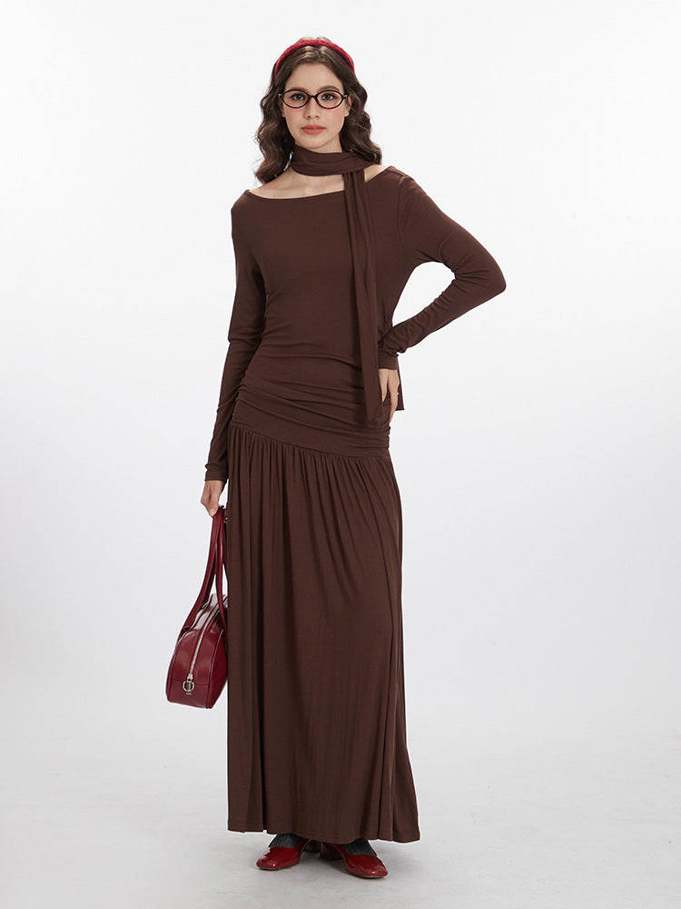 Full-length view of brown jersey maxi dress showing off-shoulder neckline, integrated scarf detail, ruched bodice, and flowing skirt, styled with burgundy handbag