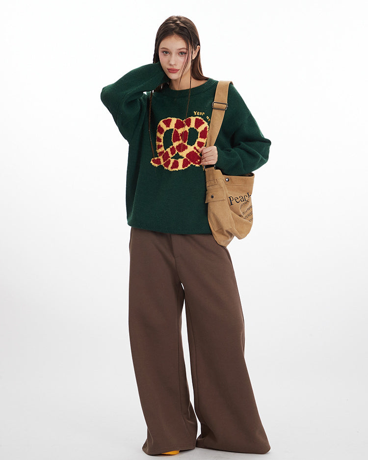 Green oversized sweater featuring red and gold pretzel graphic, styled with brown wide-leg pants and tan canvas bag