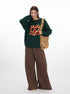 Green oversized sweater featuring red and gold pretzel graphic, styled with brown wide-leg pants and tan canvas bag