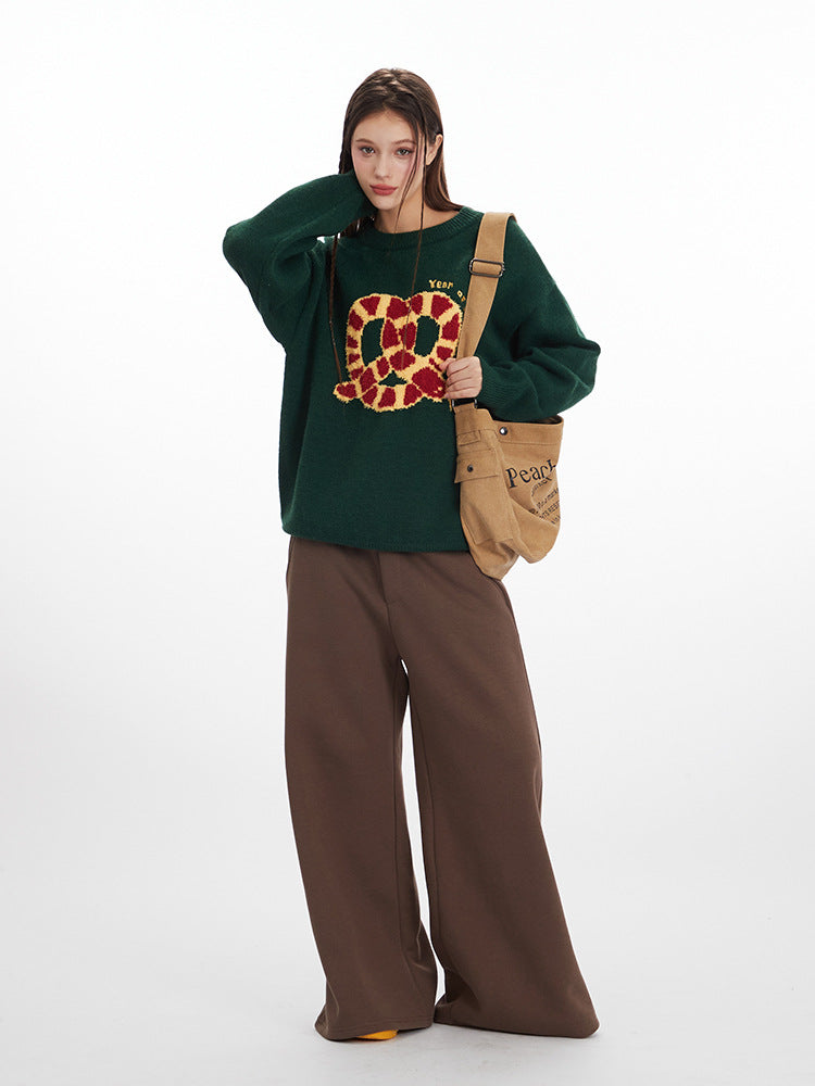 Green oversized sweater featuring red and gold pretzel graphic, styled with brown wide-leg pants and tan canvas bag