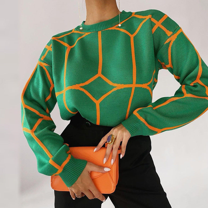 A model is wearing a green knit sweater with orange geometric pattern, black pants, and holding an orange handbag against white background