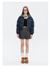 A woman is wearing a navy puffer jacket with a plaid collar, a black crop top, a gray pleated skirt, and brown platform boots