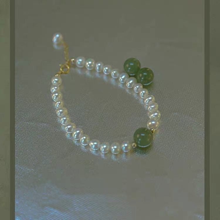 Pearl bracelet laid flat on beige fabric, featuring three jade green beads and gold-tone clasp, full view of design