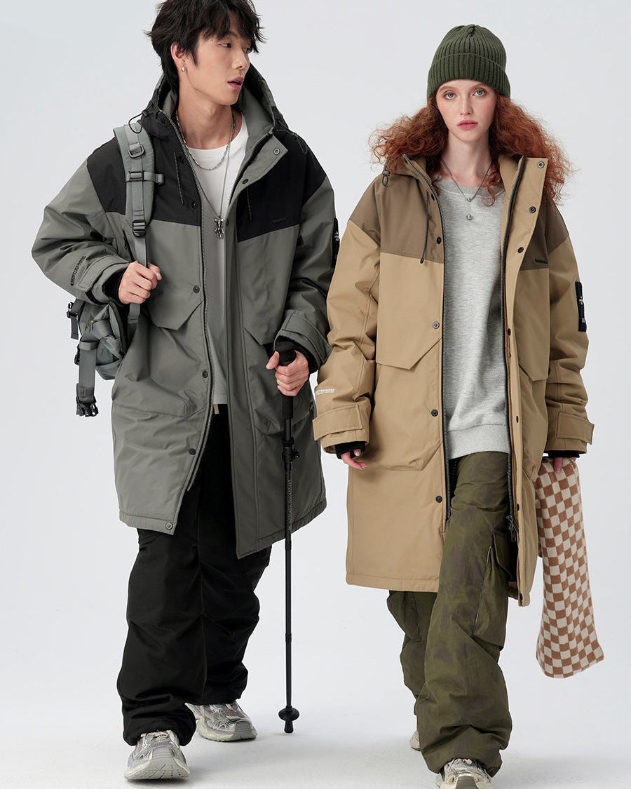 A man and woman wearing coordinating technical parkas stand against white background - his in gray/black with backpack, hers in beige/olive