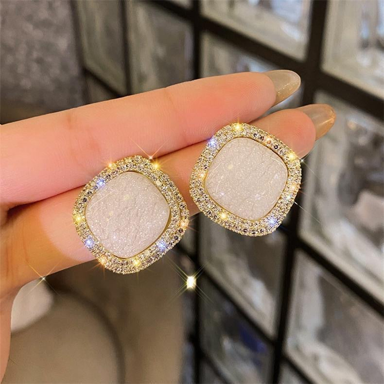 White druzy square stud earrings with sparkling crystal halos, held in hand against dark background