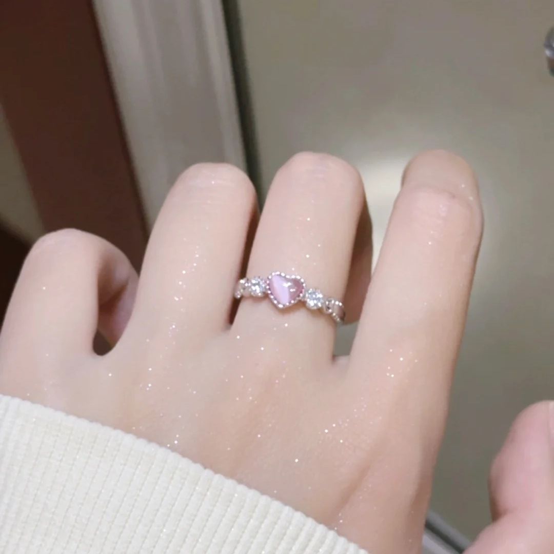 A person wearing silver promise ring with pink heart-shaped stone and crystal accents on ring finger, while wearing white sleeve