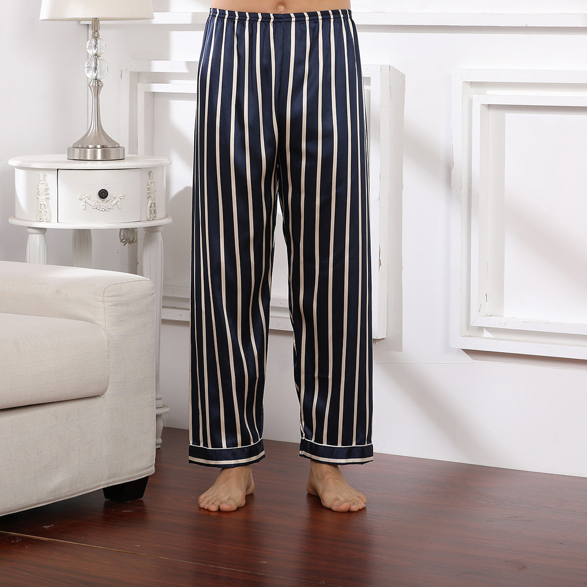 Model wearing navy blue and white vertical striped silk pajama pants with elastic waistband standing on wooden floor next to white nightstand and cream sofa