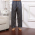 Model wearing navy blue and white vertical striped silk pajama pants with elastic waistband standing on wooden floor next to white nightstand and cream sofa