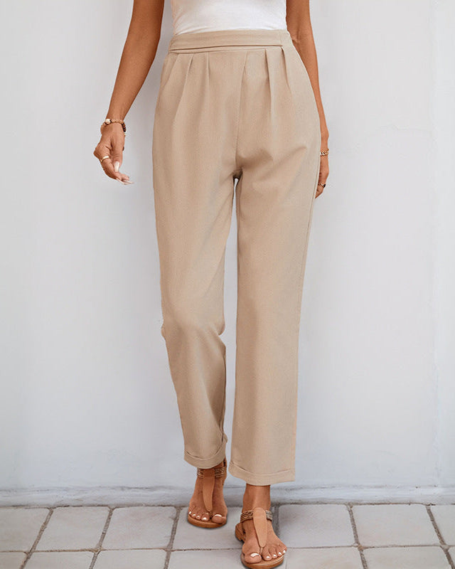  A woman wearing high-waisted beige pleated trousers with a white top and tan sandals, posing against a white wall.