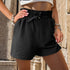Close-up of black shorts showing gathered waistband with silver D-ring belt detail and textured fabric, styled with jewelry