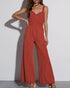 A woman wearing a rust-colored wide-leg jumpsuit with ruched detailing and adjustable straps, posing against a white wall.