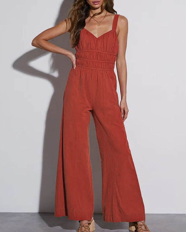 A woman wearing a rust-colored wide-leg jumpsuit with ruched detailing and adjustable straps, posing against a white wall.