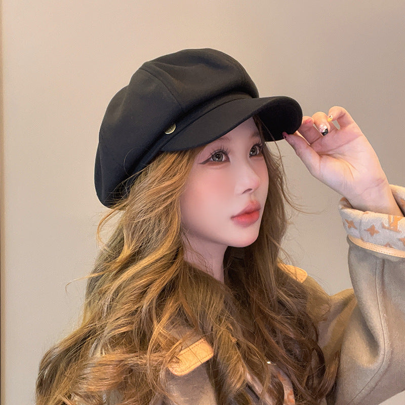 Woman in black newsboy cap touching brim, with caramel brown hair and beige outfit against neutral wall