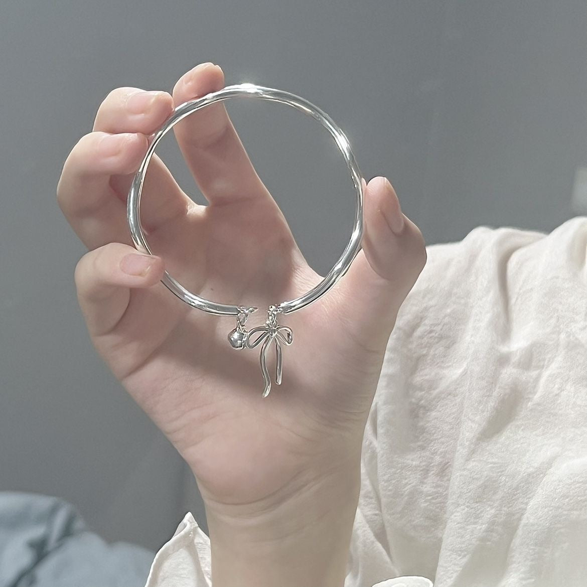 A person holding a silver bangle with bow and bell charms, wearing white clothing against a gray background