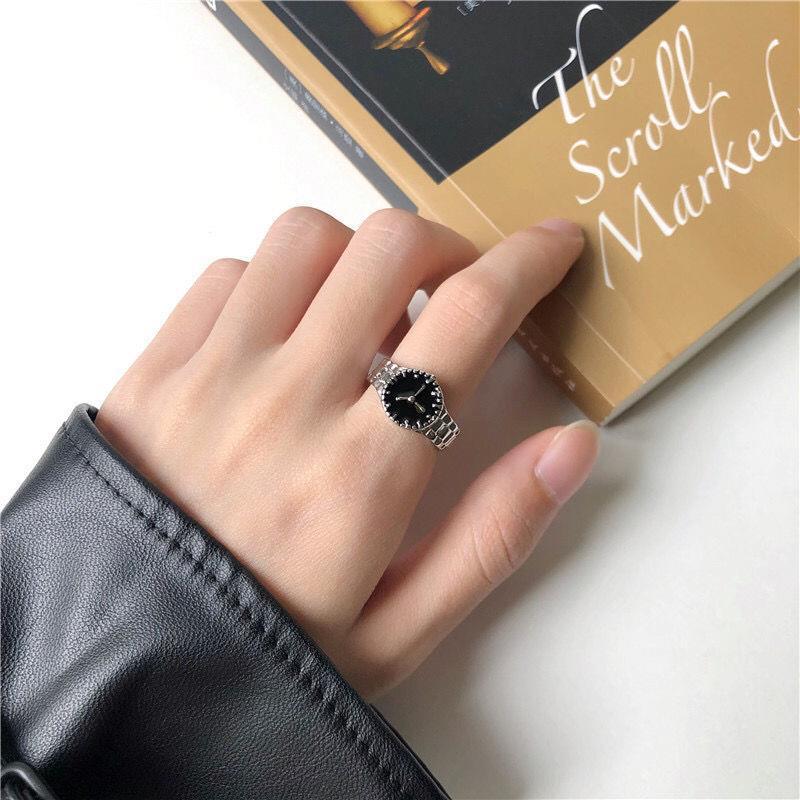 A woman's hand in black leather sleeve wearing silver watch-face ring with dark dial, resting on book with gold cover