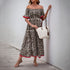 Woman in an off-shoulder leopard print maxi dress with red trim, holding a white woven tote bag, wearing sandals and jewelry.