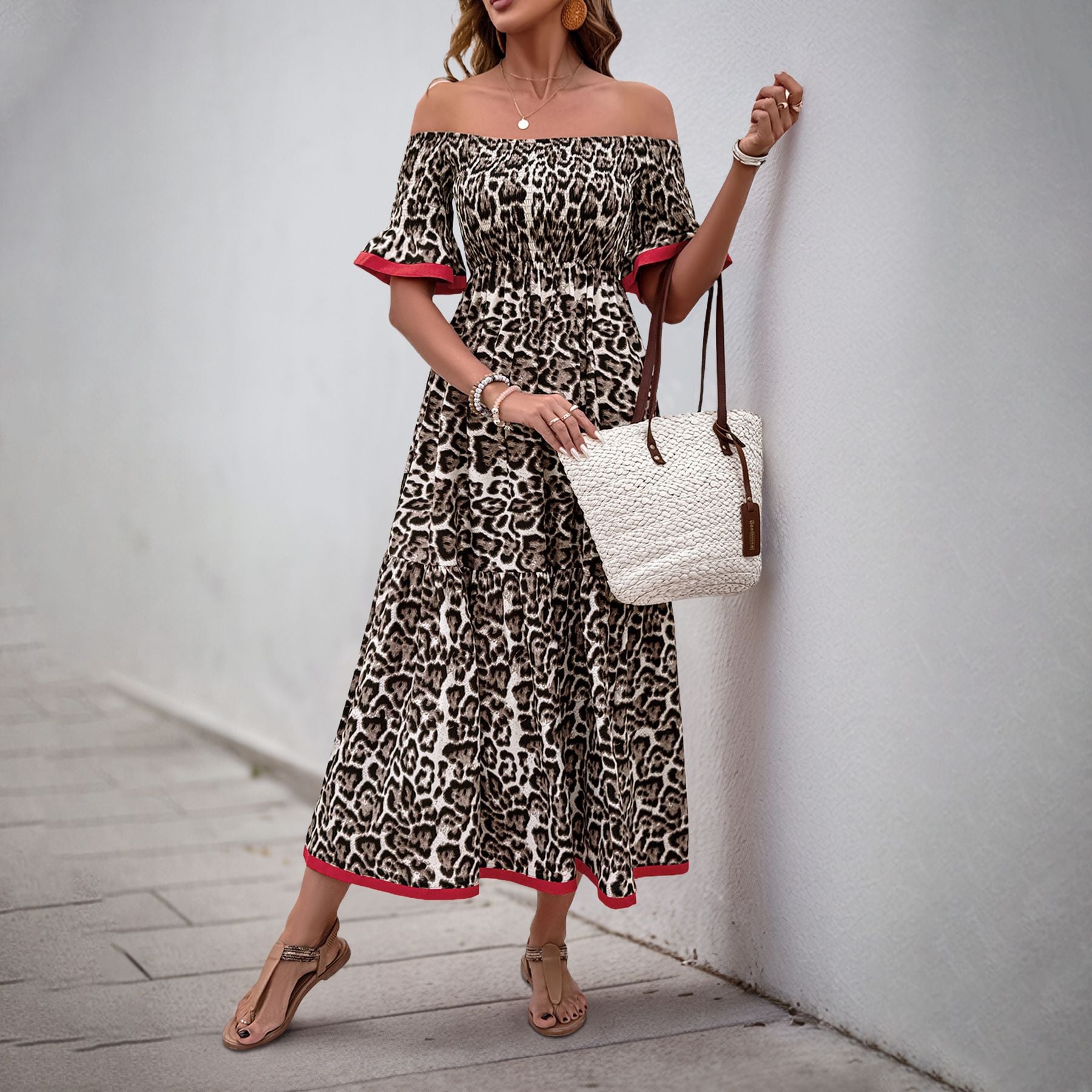 Woman in an off-shoulder leopard print maxi dress with red trim, holding a white woven tote bag, wearing sandals and jewelry.