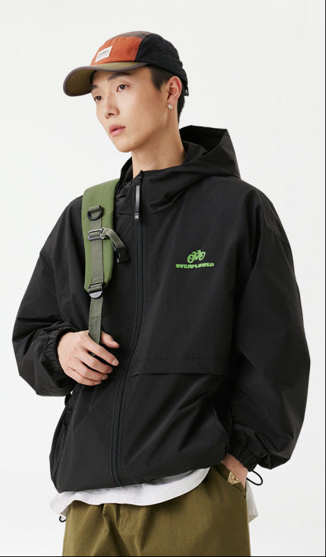A man wearing black zip-up windbreaker jacket with green bicycle logo, orange baseball cap and green shoulder bag against white backdrop