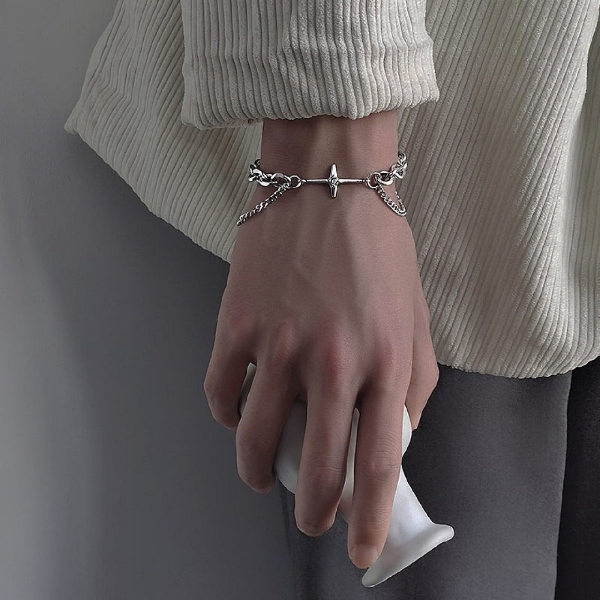 A person wearing a silver chain bracelet with a cross pendant, dressed in a white ribbed sweater, holding a white object.