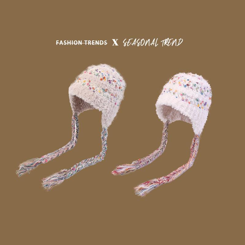 Close-up of white fuzzy winter hats with colorful speckles and multicolored braided earflaps against brown background