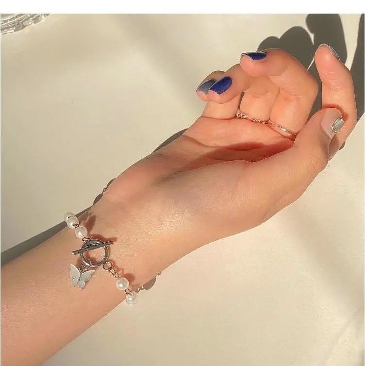 A woman wearing a pearl and silver bracelet with butterfly charm on her wrist, showing blue nails and delicate rings