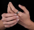 A person wearing a textured silver ring with an organic, rugged design on their finger, against a dark background.
