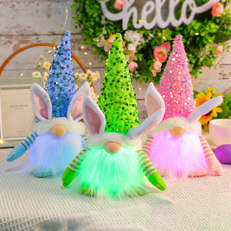 Three LED bunny gnome plush toys with sequin hats in green, pink, and blue, glowing softly on a decorated table.
