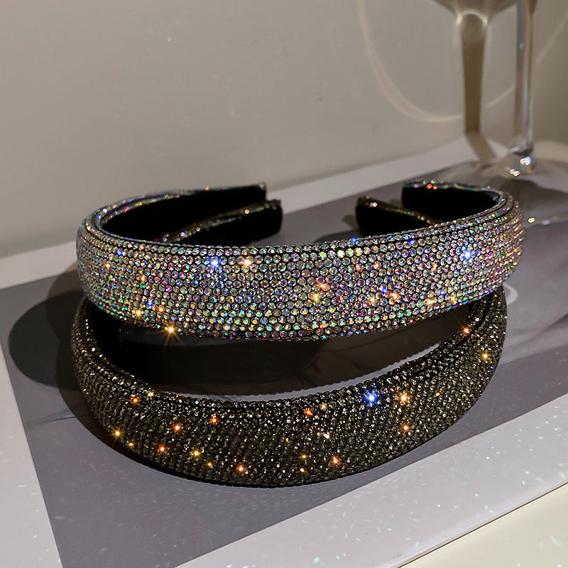 Wide black headband fully covered in iridescent rhinestones, reflecting light for a dazzling effect, placed on a glossy surface.